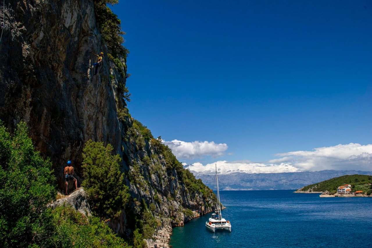 Rock climbing on a coastal cliff in the Dalmatian islands of Croatia with a luxury sailing catamaran anchored in the turquoise Adriatic Sea below.
