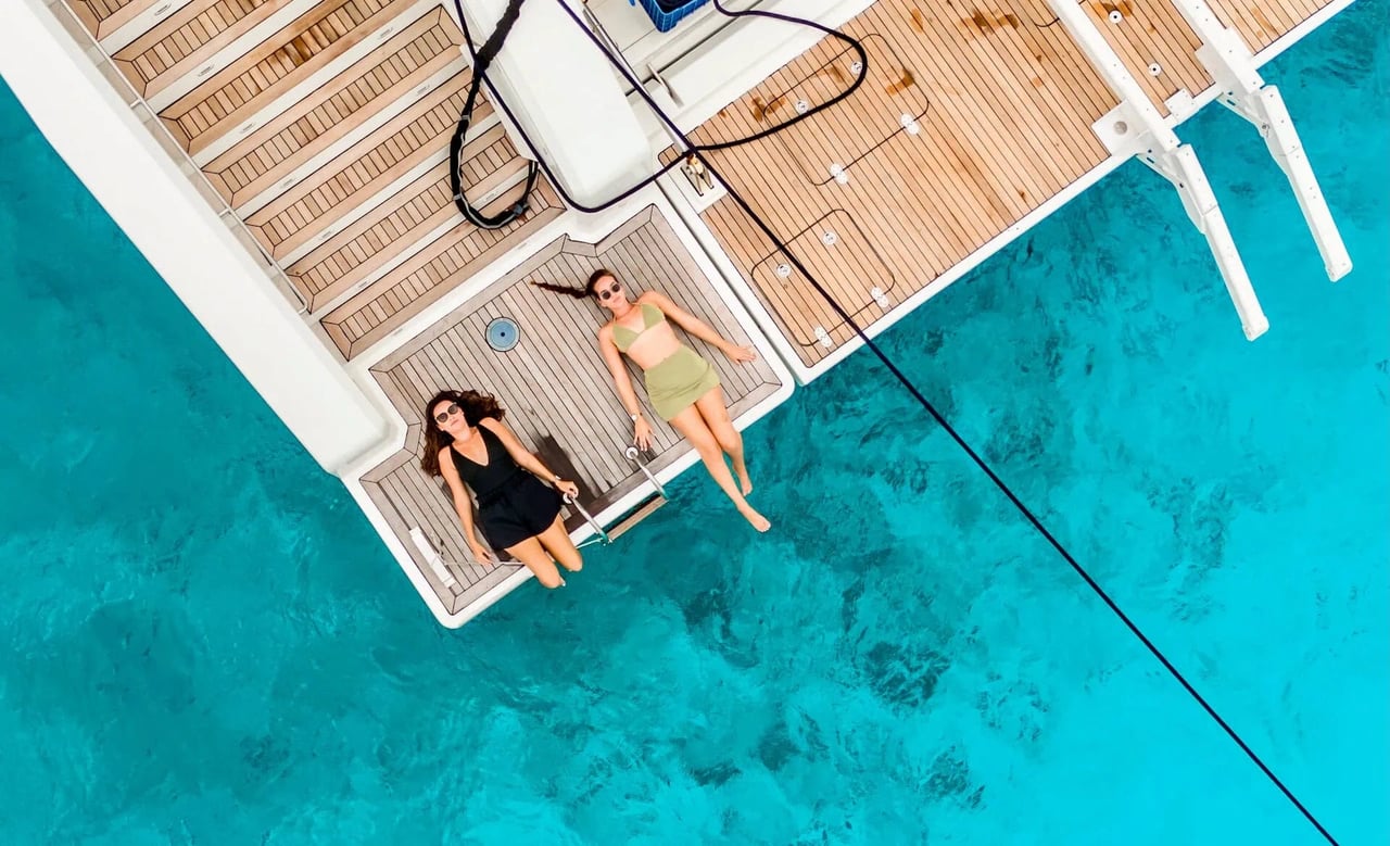 Aerial view of two women relaxing on the swim platform of a luxury sailing catamaran above crystal-clear turquoise Adriatic Sea, enjoying a peaceful summer moment while swimming and sunbathing.